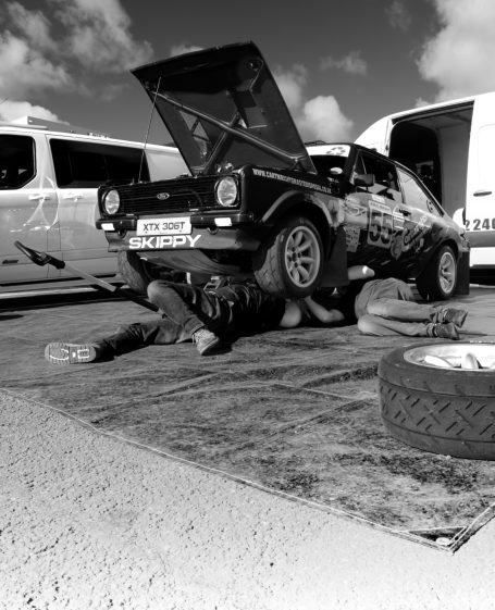 TLC for Skippy A rally car is being repaired by two mechanics under its raised chassis, by Denis Taylor