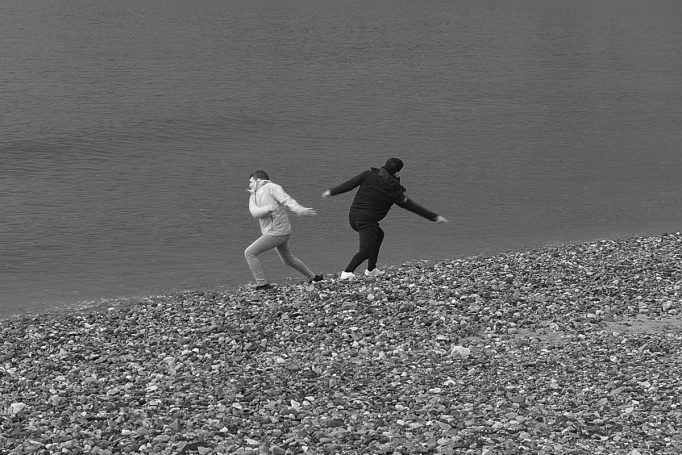 Stone Dancers Two males looking like they are doing the twist, but are really skimming stones at the sea shore, by Denis Taylor